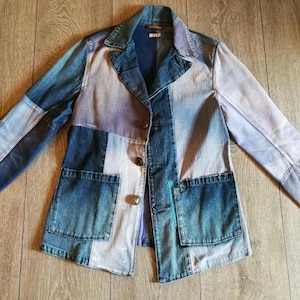Puede incluir: Chaqueta vaquera patchwork con cuello, dos bolsillos delanteros y cierre de botones. La chaqueta presenta una mezcla de paneles vaqueros azules, celeste y grises. La chaqueta está tendida sobre una superficie de madera.