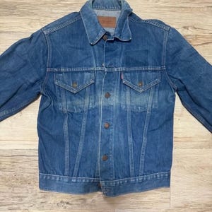 Peut inclure: Une veste en jean bleu délavé moyen avec un col pointu, une fermeture boutonnée sur le devant et deux poches poitrine à rabat boutonné. Une petite étiquette rouge est visible sur la poche gauche et une étiquette en cuir est au col.