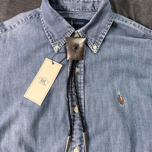 May include: A light blue denim button-down shirt with a Ralph Lauren logo and a bolo tie featuring a silver-toned, sunburst-patterned metal slide with a central stone, black braided leather cords, and silver-toned metal tips, including a RRL tag.
