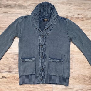 Puede incluir: Un cárdigan azul grisáceo con cuello chal, cierre de botones y dos bolsillos delanteros. El suéter tiene puños y dobladillo acanalados. La tela parece ser un tejido de punto suave. La prenda está tendida sobre una superficie de madera.