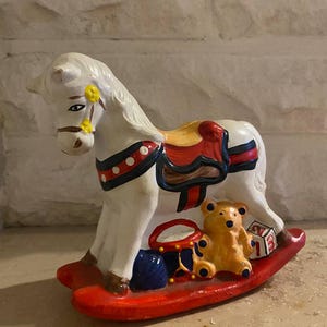 May include: A white ceramic rocking horse with a red saddle, blue and white polka dots, and yellow flowers. The horse is rocking on a red base with a teddy bear, a drum, and a toy block.