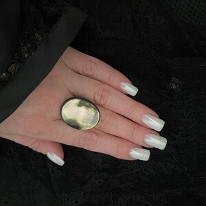 May include: An oval-shaped ring with a mother-of-pearl center, set against a dark background. The ring's iridescent surface reflects light, showcasing a range of colors. The ring is a statement piece of jewelry.