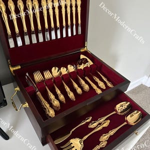 May include: A wooden box with a red velvet interior holds a set of gold-plated silverware. The box has a drawer that contains additional pieces of silverware. The silverware is ornate and features a floral design.