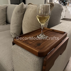 May include: A wooden tray with a hinged arm that attaches to a couch armrest. The tray is made of dark wood and has a natural finish. A glass of white wine sits on the tray. The text "DecorModernCrafts" is visible on the tray.