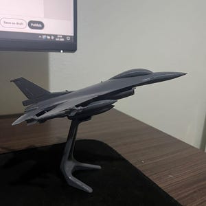 May include: A grey model fighter jet is mounted on a grey stand. The jet features a streamlined design with wings and a pointed nose. The stand has a curved, three-legged base.