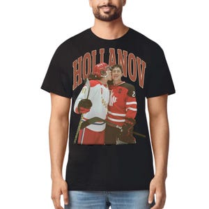 Puede incluir: Camiseta negra con una imagen retro de dos jugadores de hockey sobre hielo en rojo y blanco. La imagen tiene el texto "HOLLANOV" en una fuente vintage. Un jugador besa al otro en la mejilla, creando un ambiente nost&aacute;lgico.