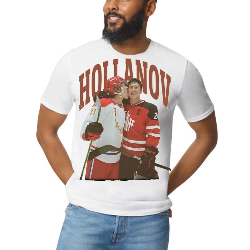 Puede incluir: Camiseta blanca con un gr&aacute;fico de dos jugadores de hockey con camisetas rojas y blancas, uno besando al otro. La palabra "HOLLANOV" est&aacute; impresa en letras marrones grandes en la parte superior. Los jugadores sostienen palos de hockey.