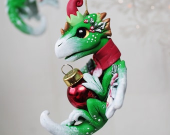 May include: A green and white dragon Christmas ornament holding a red bauble. The dragon has a red scarf and hat, with gold accents. The ornament is hanging from a gold string on a white branch.