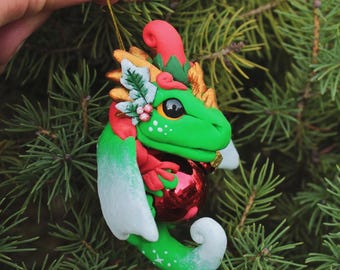 May include: Green and red Christmas ornament shaped like a dragon holding a red ornament. The dragon has gold accents and white wings.