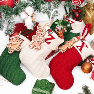 May include: Three cable-knit Christmas stockings in green, cream, and red, each featuring a white cuff. The stockings are adorned with wooden gingerbread person ornaments, with the names Yvonne and others engraved, and red embroidered letters.