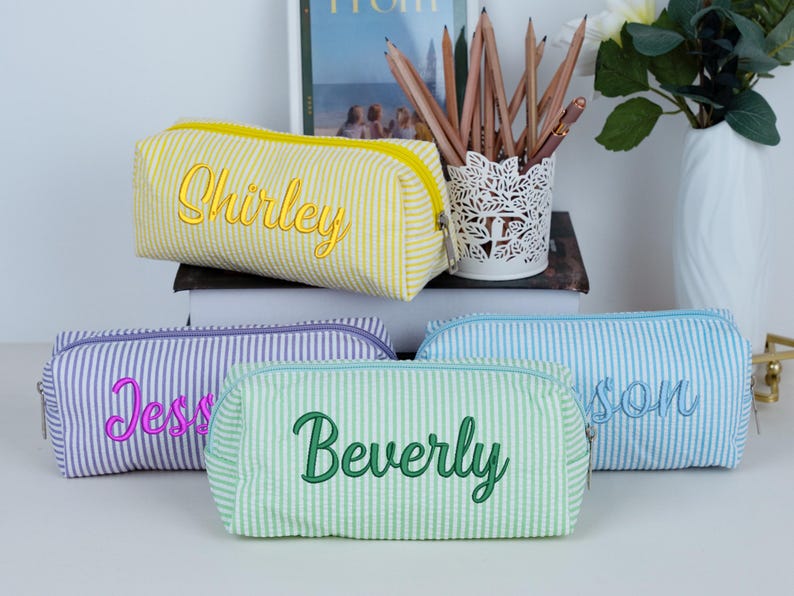 May include: Four personalized pencil cases in yellow, purple, green, and blue seersucker fabric. Each case has a different name embroidered in a contrasting color: "Shirley", "Jess", "Beverly", and "Jason". The cases have a zipper closure.