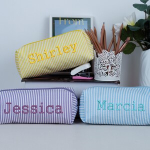 May include: Three personalized pencil cases with embroidered names. The cases are rectangular with a zipper closure and a seersucker texture. One is yellow with "Shirley", one is purple with "Jessica", and one is light blue with "Marcia".