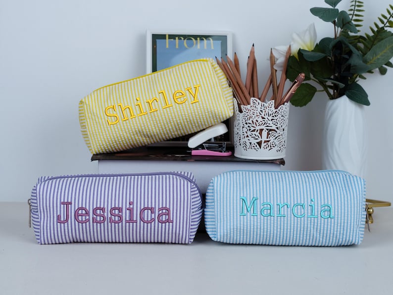 May include: Three personalized pencil cases with embroidered names. The cases are rectangular with a zipper closure and a seersucker texture. One is yellow with "Shirley", one is purple with "Jessica", and one is light blue with "Marcia".