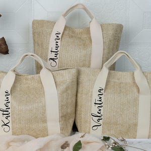 K&ouml;nnte beinhalten: Drei beigefarbene gewebte Tragetaschen mit cremefarbenen Griffen und Tr&auml;gern. Die Taschen tragen Namen auf den Tr&auml;gern: "Autumn", "Katherine" und "Valentina" in Schreibschrift. Die Taschen sind auf einer wei&szlig;en Oberfl&auml;che angeordnet.
