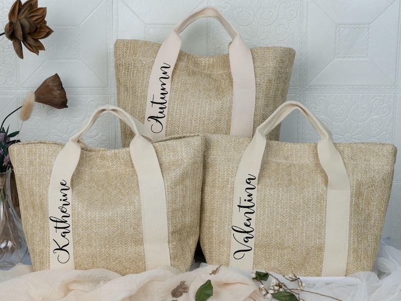 K&ouml;nnte beinhalten: Drei beigefarbene gewebte Tragetaschen mit cremefarbenen Griffen und Tr&auml;gern. Die Taschen tragen Namen auf den Tr&auml;gern: "Autumn", "Katherine" und "Valentina" in Schreibschrift. Die Taschen sind auf einer wei&szlig;en Oberfl&auml;che angeordnet.