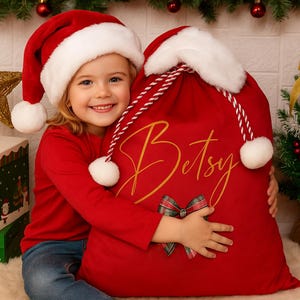 May include: A child in a Santa hat and red top embraces a large red Christmas sack. The sack is personalised with the name "Betsy" in gold script and a tartan bow, festive decorations in the background.