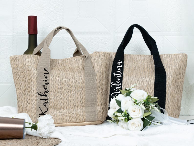 K&ouml;nnte beinhalten: Zwei gewebte Tragetaschen mit langen Henkeln. Eine hat einen beigen Henkel mit dem Namen "Katherine" in schwarzer Schrift, die andere hat einen schwarzen Henkel mit dem Namen "Valentina". Eine Weinflasche und ein Strau&szlig; wei&szlig;er Blumen sind davor.