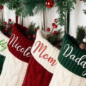Könnte beinhalten: Vier Weihnachtsstrümpfe hängen an einer dekorativen Girlande. Die Strümpfe sind aus Zopfstrick in Grün, Rot und Weiß gefertigt und mit Namen in passenden Farben bestickt: "Daddy", "Nicole", "Mom" und "Daddy".