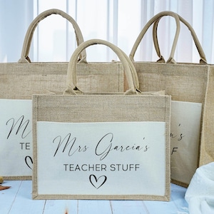 Può includere: Tre borse tote in iuta beige con tasche color crema. La borsa centrale presenta la scritta "Mrs. Garcia's TEACHER STUFF" con un simbolo a cuore. Le altre borse hanno la lettera "M" e la parola "TEACHER". Ogni borsa ha un manico beige abbinato.