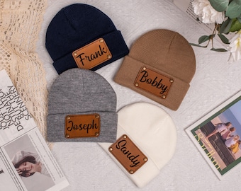 May include: Four personalised beanies in navy blue, brown, grey, and white. Each beanie has a brown leather patch with a name in black script: Frank, Bobby, Joseph, and Sandy. The beanies are on a textured surface.