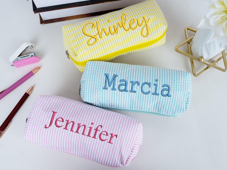 May include: Three personalized pencil cases in yellow, light blue, and pink, each with a name embroidered in a contrasting color. The yellow case says "Shirley", the light blue says "Marcia", and the pink says "Jennifer".