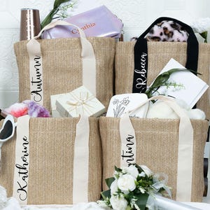 K&ouml;nnte beinhalten: Jute-Tragetaschen mit personalisierten Namen auf den Tr&auml;gern. Die Taschen sind mit Geschenken gef&uuml;llt, darunter eine Weinflasche, Sonnenbrille und eine Geschenkbox. Die Taschen haben cremefarbene Tr&auml;ger und sind mit Blumen dekoriert.