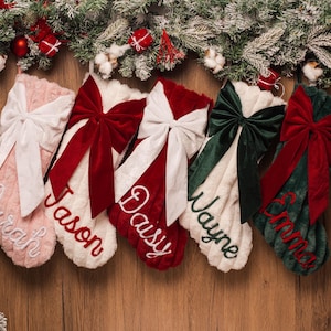 Könnte beinhalten: Fünf weiche Weihnachtsstrümpfe aus Kunstfell, jeweils mit einer großen Samtschleife und personalisierter Stickerei versehen. Die Strümpfe sind in Hellrosa, Weiß, Rot, Creme und Dunkelgrün gehalten und tragen die Namen Sarah, Jason, Daisy, Wayne und Emma.