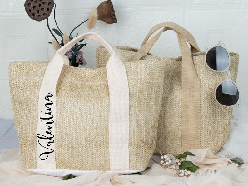 K&ouml;nnte beinhalten: Zwei gewebte Stroh-Tragetaschen mit cremefarbenen Griffen und Akzenten. Eine Tasche hat den Namen "Valentina" vertikal in schwarzer Schrift aufgedruckt. Sonnenbrillen liegen auf der Seite einer Tasche. Die Taschen stehen auf einer wei&szlig;en Oberfl&auml;che.