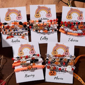 Op de afbeelding: Vijf sets kralenarmbanden op witte kaarten, elk met een regenboog- en pompoenafbeelding en de tekst Happy Halloween. De armbanden bevatten oranje, zwarte en witte kralen met bedels in de vorm van pompoenen, vleermuizen en spoken.