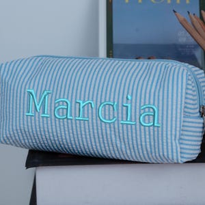 May include: A blue and white striped pencil case with the name "Marcia" embroidered in light blue. The rectangular case has a zipper closure and is sitting on a dark surface. Pencils are visible in the background.