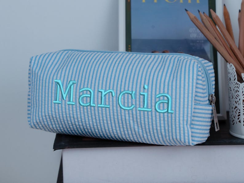 May include: A blue and white striped pencil case with the name "Marcia" embroidered in light blue. The rectangular case has a zipper closure and is sitting on a dark surface. Pencils are visible in the background.