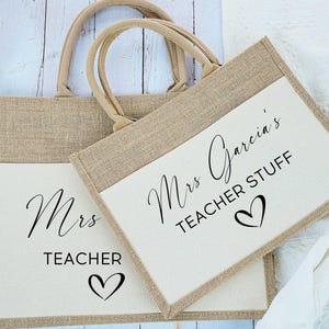 May include: Two beige burlap tote bags with cream-colored pockets. Each bag has a set of matching handles. The text "Mrs. Garcia's TEACHER STUFF" and a heart are printed on the front of the bags.