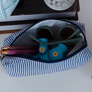 May include: A blue and white striped zippered pouch, open to reveal sunglasses, pens, and a blue toy. The pouch is made of fabric and has a navy blue zipper. The sunglasses have a green frame and brown lenses.