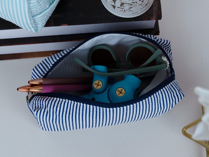 May include: A blue and white striped zippered pouch, open to reveal sunglasses, pens, and a blue toy. The pouch is made of fabric and has a navy blue zipper. The sunglasses have a green frame and brown lenses.