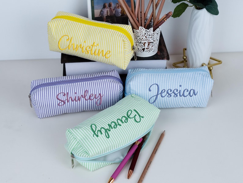May include: Four personalized pencil cases in yellow, purple, blue, and green seersucker fabric with white stripes. Each case has a different name embroidered in a matching color: "Christine", "Shirley", "Jessica", and "Beverly".