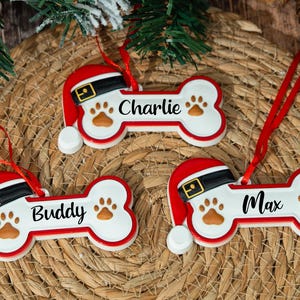 May include: Three Christmas ornaments shaped like dog bones, each with a Santa hat. The ornaments are white with red trim and feature paw prints and the names "Charlie", "Buddy", and "Max". A black belt and gold buckle detail the Santa hat.