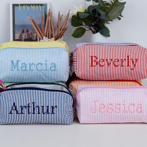 May include: A collection of personalized pencil cases in various colors, including yellow, blue, red, orange, and pink. Each case features a vertical striped pattern and a name embroidered in a contrasting color. The cases are zippered and appear to be made of fabric.