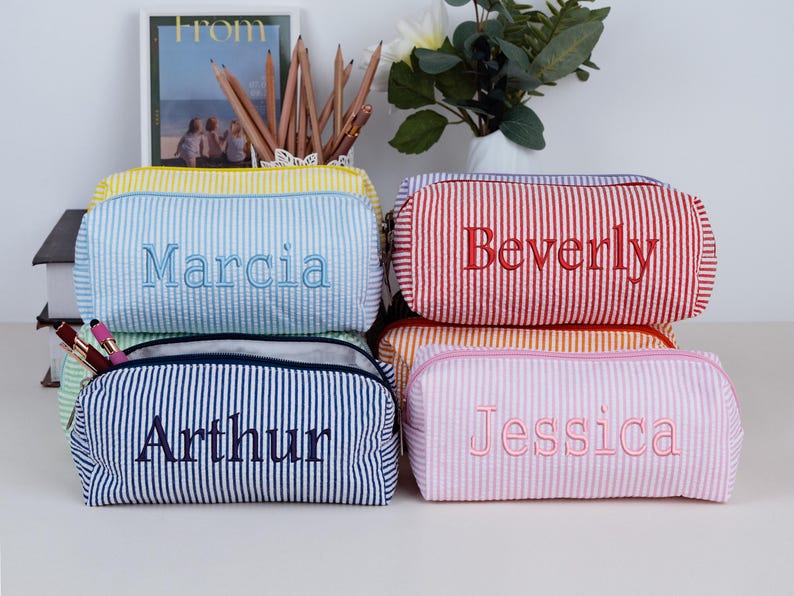 May include: A collection of personalized pencil cases in various colors, including yellow, blue, red, orange, and pink. Each case features a vertical striped pattern and a name embroidered in a contrasting color. The cases are zippered and appear to be made of fabric.