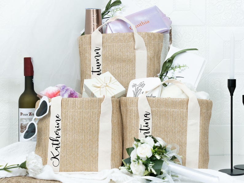 K&ouml;nnte beinhalten: Drei gewebte Tragetaschen mit wei&szlig;en Bandgriffen, jede mit einem auf dem Band aufgedruckten Namen. Die Taschen sind mit Geschenken gef&uuml;llt, darunter eine Weinflasche, eine Sonnenbrille und ein kleiner Strau&szlig; wei&szlig;er Rosen.