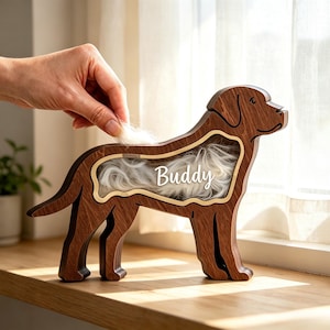 May include: A dark brown wooden dog-shaped display frame with a hollow center filled with white fur. The name Buddy is printed in white script across the center of the frame. The frame is designed to hold pet fur as a keepsake memorial.
