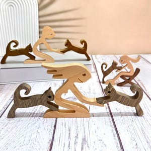 Puede incluir: Figuras de madera de personas y gatos. Las figuras están en varias poses, incluyendo una mujer sosteniendo un gato, un hombre sosteniendo un gato y un gato saltando. Las figuras están hechas de madera marrón claro y tienen un acabado natural.