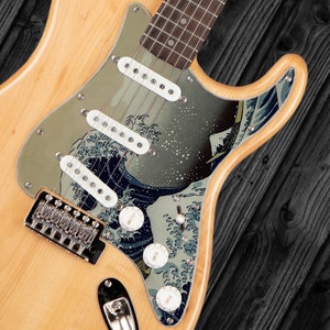 May include: A natural wood electric guitar with a custom pickguard featuring a Japanese wave design. The pickguard is silver with a blue and white wave pattern. The guitar has three knobs and a chrome bridge.