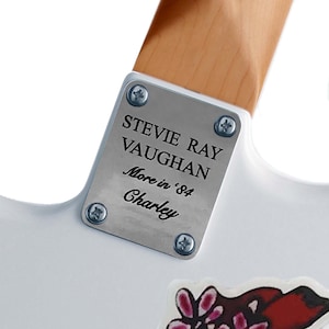 May include: A silver metal plate with the text "Stevie Ray Vaughan More in '84 Charley" on a white guitar.