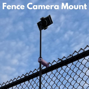May include: A black metal fence camera mount with a pink plastic clamp. A black smartphone is attached to the mount, facing upwards.
