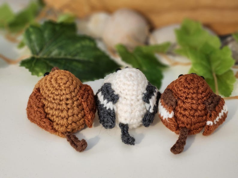Puede incluir: Tres adornos de p&aacute;jaros hechos a mano con ganchillo en tonos marr&oacute;n, blanco y gris. Los p&aacute;jaros tienen peque&ntilde;os ojos negros y una cola. El fondo incluye hojas verdes y un fondo neutro borroso.