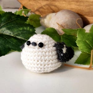 Puede incluir: Un p&aacute;jaro de ganchillo blanco hecho a mano con ojos negros y alas grises y negras. El p&aacute;jaro es redondo y esponjoso, con un dise&ntilde;o simple y encantador. Hojas verdes y un fondo de color claro proporcionan un entorno natural.