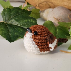 Puede incluir: Un p&aacute;jaro de ganchillo hecho a mano con hilo marr&oacute;n y blanco. El p&aacute;jaro tiene ojos de cuentas negras y una peque&ntilde;a cola marr&oacute;n. Se coloca sobre un fondo de hojas verdes y una superficie de color neutro.
