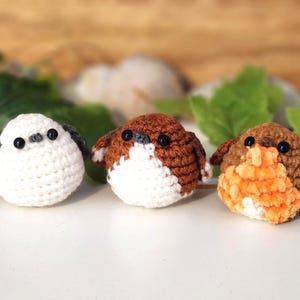 Puede incluir: Tres adornos de p&aacute;jaros hechos a mano con ganchillo. Uno es blanco con detalles grises, los otros son marrones y naranjas. Cada p&aacute;jaro tiene ojos negros y est&aacute; sobre una superficie blanca. Se ven hojas verdes y un fondo de madera.