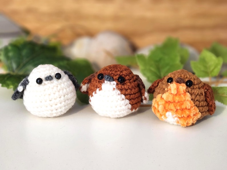 Puede incluir: Tres adornos de p&aacute;jaros hechos a mano con ganchillo. Uno es blanco con detalles grises, los otros son marrones y naranjas. Cada p&aacute;jaro tiene ojos negros y est&aacute; sobre una superficie blanca. Se ven hojas verdes y un fondo de madera.