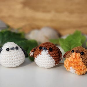 Puede incluir: Tres figuras de p&aacute;jaros de ganchillo hechas a mano. Los p&aacute;jaros son blancos, marrones y naranjas, con ojos negros. Son peque&ntilde;os y redondos, y parecen estar sobre una superficie blanca. Hay hojas verdes en el fondo.